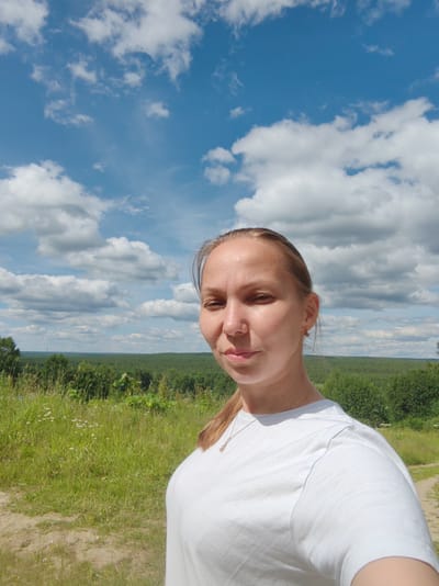 In the city of Chukhlem, Russia, a young woman with blonde hair pulled back poses for a selfie outdoors under a bright blue sky with scattered white clouds. She is wearing a white t-shirt and a delicate necklace. The background features a lush green landscape with a forest in the distance and a dirt path to her right. The lighting suggests it is daytime, likely on a sunny afternoon.