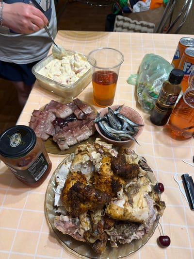 A high-angle, close-up shot shows a table laden with food, likely a meal. The table is covered with a light-colored checkered tablecloth. In the center of the frame, a platter holds a partially eaten roasted chicken, its skin dark and crispy in places. To the left of the chicken, a small bowl contains a pile of small, silver fish, possibly sardines or anchovies. Adjacent to the fish is a dark brown platter with slices of what appears to be pickled herring or other cured fish, with a dark, shiny glaze.

Further left, a clear glass container holds a creamy, yellowish salad, possibly potato salad or coleslaw, with chunks of red and white ingredients visible. A spoon is positioned in the salad, and a person's hands and arm are visible on the lower left, reaching for the salad with the spoon. A clear glass with an amber-colored liquid, likely beer or iced tea, stands near the salad.

In the background, two cans of orange soda, one labeled "FA," are visible. A small bottle with a dark liquid and a label in Cyrillic script, possibly soy sauce or a seasoning, is also present. A black lighter with "Cricket" printed on it lies near the top of the frame. Two dark red cherries are placed as a garnish on the chicken platter. The scene suggests a casual meal or gathering, possibly in a home setting. The lighting indicates it could be daytime. The text visible includes brand names like "FA" and "Cricket," as well as Cyrillic text on a bottle, which includes "Руна" and "ТЕРІЯК."
