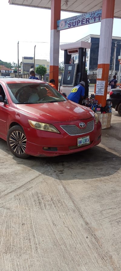 The image depicts an active scene at a gas station identified as "F & L SUPER 11" in Akure, Nigeria. A red Toyota Camry, license plate "ABUJA BWR-138CK", is parked on a rough concrete surface in the foreground, facing slightly left. A gas station attendant, wearing a blue shirt with "SUPER 69" on the back, is positioned at a black and white fuel dispenser, actively fueling the red vehicle. The car's driver is partially visible through the windshield. To the right of the fuel dispenser, a small makeshift retail display is set up on a tiled base, offering various items like sunglasses and hats. A pillar adjacent to this display bears a white sign listing "FARDAS" and multiple Nigerian bank account details. In the background, other individuals are present, including a person in an orange shirt near the fuel pump and another in a blue shirt further right, near a partially visible black vehicle. Beyond the gas station structure, a building marked "HEXIM BANK" is visible, along with other urban structures and sparse vegetation under a bright sky.