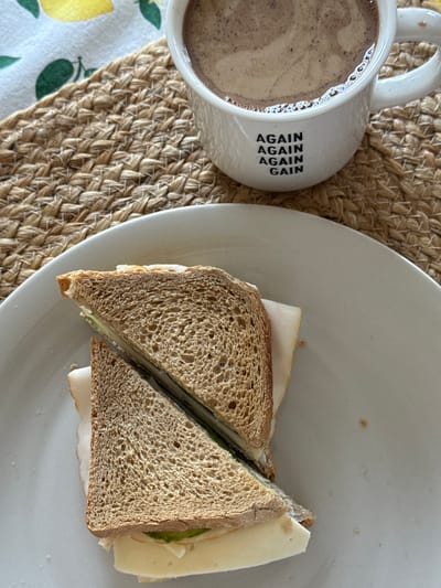 A top-down shot shows a mug of coffee next to a sandwich cut in half on a white plate, resting on a woven mat. A tablecloth with a lemon pattern is visible in the lower left corner. The mug is white with the words "AGAIN AGAIN AGAIN" printed vertically. The sandwich appears to be made with whole wheat bread, cheese, sliced turkey, and green vegetables. The location is not identifiable from the image.