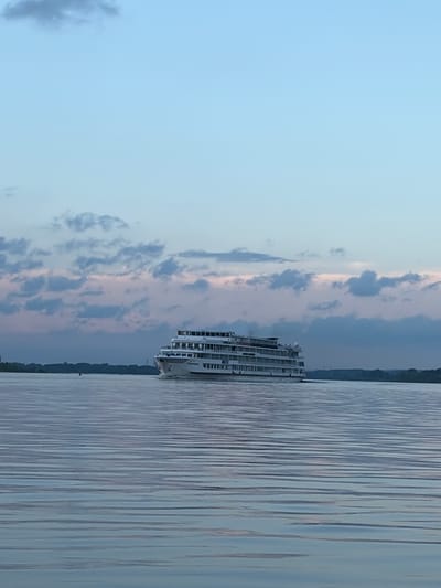 A large, multi-decked white cruise ship is sailing on a wide, calm river under a pale blue sky with scattered pink and grey clouds. The ship is positioned on the right side of the frame, moving towards the left. The water's surface has numerous horizontal ripples. The horizon shows a dark, tree-lined shore with a small red buoy visible in the distance on the left. The image captures a serene moment, likely at dusk or dawn, on a river in or near the city of Olkhovka, Russia. There are no people visible in the image.
