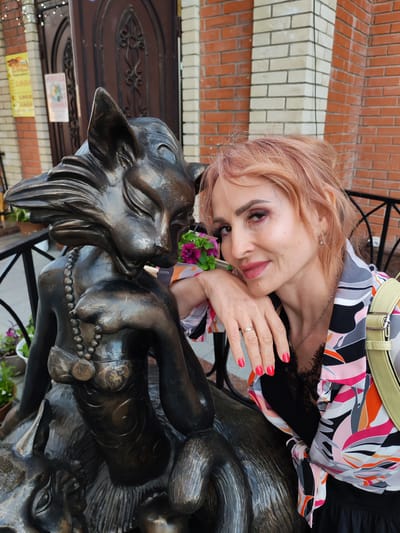 The image shows a woman posing next to a bronze statue of a cat. The statue is of a female cat, sitting and wearing a beaded necklace and a brassiere-like garment. The woman, who has short, pinkish hair and is wearing a colorful, patterned blouse and a black dress, leans her head on her hand, resting it on the statue. Her left hand, with bright red nail polish, is visible, showing a ring on her ring finger. She is looking towards the camera with a slight smile. The background suggests an outdoor setting, with a brick building and some potted plants visible. There are also posters on the wall behind the statue, and a decorative wrought iron fence. This scene is located in Yoshkar-Ola, Russia, specifically at the cat monument on Bulvar Chaynoy.