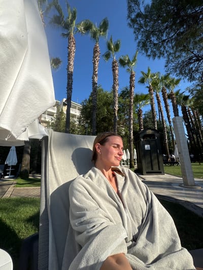 A woman is relaxing on a lounge chair on a sunny day, wrapped in a grey robe. She is positioned in the foreground, with her eyes closed and her face tilted towards the sun. Behind her, there are tall palm trees, a white building, and a shower structure. The scene appears to be a resort or hotel area in Manavgat, Turkey, given the presence of palm trees, resort-style architecture, and outdoor shower facilities. The ground is covered with green grass and a paved walkway. The overall impression is one of leisure and relaxation in a warm, sunny climate.