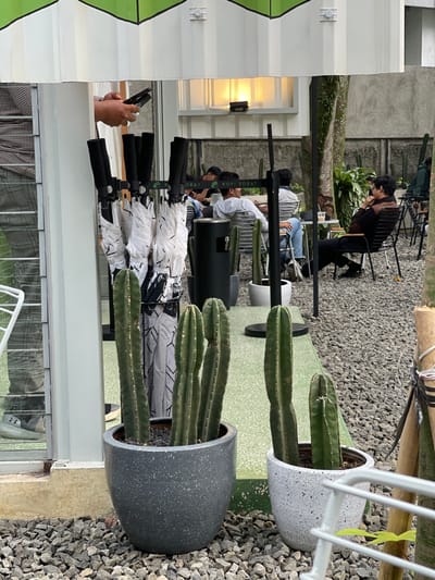 The image depicts an outdoor cafe area, identified as a "Kopi Sobat" establishment, located in Bogor, Indonesia. The ground is covered uniformly with light grey gravel.
In the foreground, two large pots containing tall, green columnar cacti are prominently positioned. The pot on the left is dark grey and speckled, holding multiple cacti, while the pot on the right is white and speckled, containing two cacti and partially obscured by a white metal railing.
The midground features a white building structure with a green, geometric-patterned roof overhang. Beneath this overhang, a stand holds numerous folded white umbrellas with black handles and black-and-white patterned fabric. Above the umbrellas, a person's hands are visible, holding a black smartphone that appears to be in a folded or flip position. To the left of the umbrella stand, a partial reflection of a person wearing plaid pants and dark shoes is visible in a glass surface.
The background reveals an outdoor seating area occupied by at least five individuals. Three people are seated at a table on the left, one wearing a light-colored hoodie and looking to the right. Two people are seated at a table on the right; one individual in a dark long-sleeved shirt and dark pants is leaning back in a black metal chair, with their right hand near their chin. Small, light-colored round tables and black metal chairs are distributed across the gravel. Additional potted cacti are interspersed within the seating area. A black sign displaying "KOPI SOBAT" in white text is visible above the seated patrons. Green foliage and trees are visible in the distant background against a grey wall, and a window on a white wall in the upper background emits a warm yellow light.