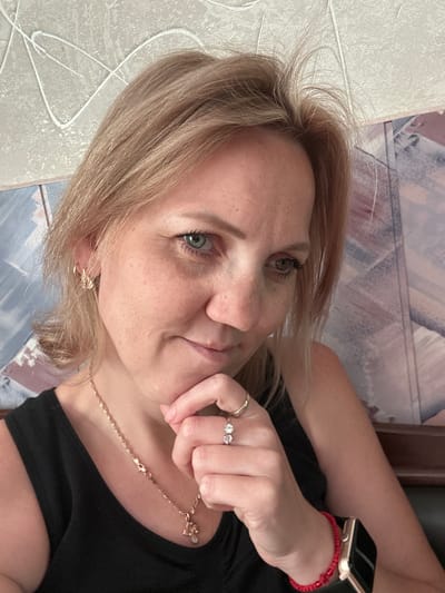 An outdoor medium shot shows a light-skinned woman with blonde hair and light blue eyes, wearing a black tank top, gold necklace, and two rings on her right hand. Her hand is resting on her chin. She has freckles across her nose and cheeks. A red beaded bracelet is visible on her left wrist, along with a smartwatch. The background consists of a textured wall and patterned wallpaper. The setting appears to be a home in Gelendzhik, Russia.