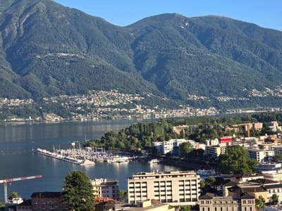 This media file depicts a scenic, sun-drenched view of Muralto, Switzerland, likely taken in the late afternoon given the warm lighting and long shadows. The scene is dominated by the majestic, tree-covered mountains that form a dramatic backdrop to the town and the expansive, calm lake. The water reflects the clear blue sky, creating a serene atmosphere.
In the foreground, the urban landscape of Muralto unfolds, characterized by a mix of modern buildings and older structures with terracotta roofs. A prominent marina juts into the lake, filled with numerous sailboats and yachts, suggesting a leisurely boating activity. A large construction crane is visible to the left of the marina, hinting at ongoing development or renovation in the area. Trees are scattered throughout the town, adding greenery to the urban setting.
The overall impression is one of peace and tranquility, with the natural beauty of the mountains and lake harmoniously coexisting with the town. The clear weather and bright sunlight contribute to a feeling of warmth and pleasantness, inviting exploration of the picturesque location. There is no visible text in the image.