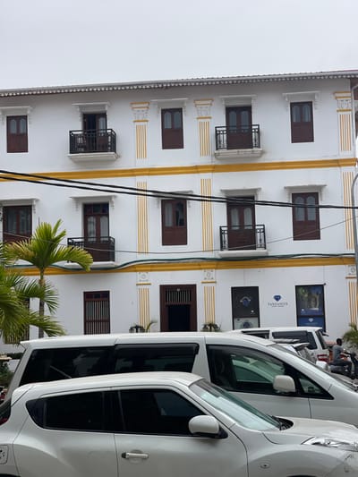 A white building with brown windows and yellow accents. Some windows have small balconies. Two vertical black wires run down the building's face. White vehicles obstruct the right side, their windows tinted dark. A small palm tree sits at the base of the building. The building's upper windows display "Tanzanite" text.
