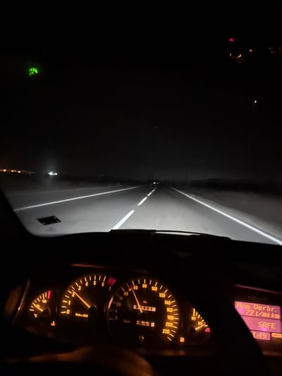 The image presents a nighttime view from the driver's perspective inside a vehicle moving along a paved road in the city of Trapoklovo, Bulgaria. The road ahead is illuminated by the vehicle's headlights, revealing distinct white dashed lane markings and solid white shoulder lines. Distant lights are visible on the left horizon, suggesting an urban or industrial area. The vehicle's dashboard is illuminated with an orange glow, prominently displaying the speedometer indicating approximately 60 km/h and the tachometer at roughly 2000-2500 RPM. The odometer reads 829726 kilometers, and the trip meter shows 0099.3 kilometers. The fuel gauge indicates approximately one-quarter tank. A digital information display on the right side of the dashboard shows "Ком. Verb. 7.7 l/100 km", "SAFE", and "1:49". Multiple warning lights are active on the instrument panel. A green light source or reflection is present in the upper left of the frame. The presence of a driver operating the vehicle is inferred from the perspective and active dashboard instrumentation.