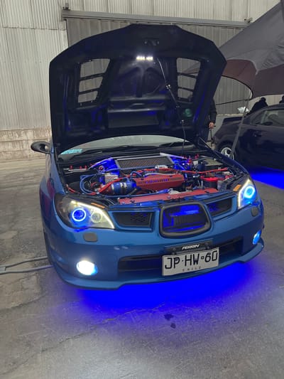 The image shows a blue Subaru Impreza with its hood open, displaying a modified engine bay with extensive blue and red lighting. The car is parked on a concrete floor in what appears to be an indoor or covered venue, with a corrugated metal wall in the background. The car is decorated with custom headlights that feature blue halo rings and underglow lighting of the same color illuminates the ground around it. A license plate with the designation "JP HW 60 CHILE" is visible on the front bumper. In the background, a dark-colored car and a person wearing dark clothing are partially visible, along with a large umbrella. The overall impression is of a car show or exhibition.