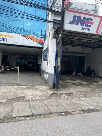 The scene is located in Kecamatan Margahayu, Indonesia, at coordinates -6.973 latitude and 107.573 longitude. It shows a street corner with a JNE Express storefront on the right. The storefront has a blue awning with white lettering and a sign that reads "JNE EXPRESS".  The building is painted white and has a number "228" on the side. To the left of the street is a building with a blue glass facade, a black car parked in front, and a sign that reads "Diri". There are power lines running across the scene, and the sidewalk is broken and cracked. There are a few motorcycles parked in front of both storefronts and a person walking away from the JNE Express storefront.  The view is taken from the street looking down the sidewalk.