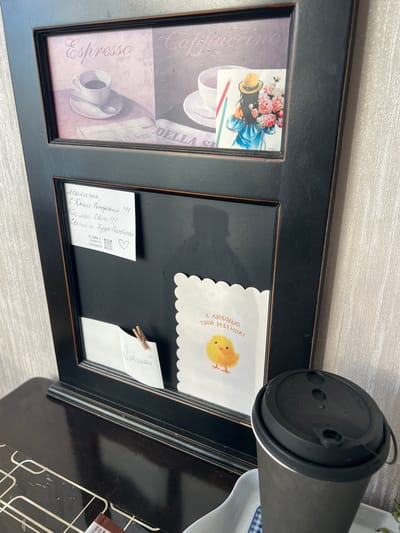 The image shows a black framed memo board with several items attached. The top section of the board features a graphic with the word "Espresso" and an illustration of a coffee cup, next to another graphic with the word "Cappuccino" and an illustration of a coffee cup with a girl holding flowers. Below this, there is a white note with handwritten text and a heart drawing, attached to the board. A small white card with a drawing of a chick and text in Russian is also attached to the board. A small piece of paper with the word "Любимой" (beloved) is clipped to the board with a small wooden clothespin. A dark-colored coffee cup with a black lid sits on a dark wooden surface in front of the memo board. The memo board is leaning against a wall with textured wallpaper. The overall scene appears to be an indoor setting, likely a home or office. The location in Barnaul, Russia is not specified in the image itself.