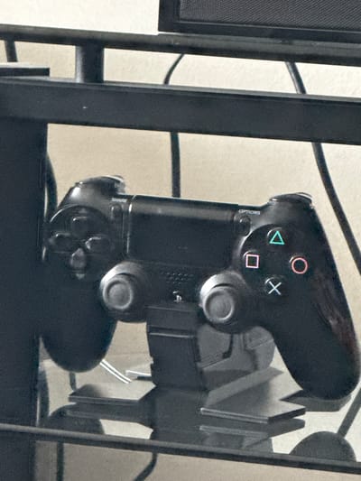 A black PlayStation 4 controller is the primary subject in this image, angled vertically and resting on a black surface. It is secured by a black metal stand, parts of which are visible framing the controller on the left and right. The controller's face buttons—a red circle, a blue triangle, a pink square, and a white cross—are clearly visible. The "OPTIONS" button is also discernible on the left side of the controller. The controller is connected to a black cable that runs out of frame upwards. The background is a plain, light-colored surface, possibly a wall, with some shadows cast by the stand and controller, suggesting the lighting is coming from the front. The image appears to be taken indoors, in a room with a neutral and somewhat dim ambiance. No people are present. The specific location is New Westminster, Canada.
