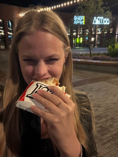 The image shows a young woman eating a shawarma outdoors at night. She has long blonde hair and is wearing a ring on her right hand. The background is blurred but appears to be a street scene with neon lights and trees. The woman is looking down at her food, and her expression is neutral. The text on the shawarma wrapper is in Russian and appears to be a label with product information. The overall impression is one of a casual, late-night meal.