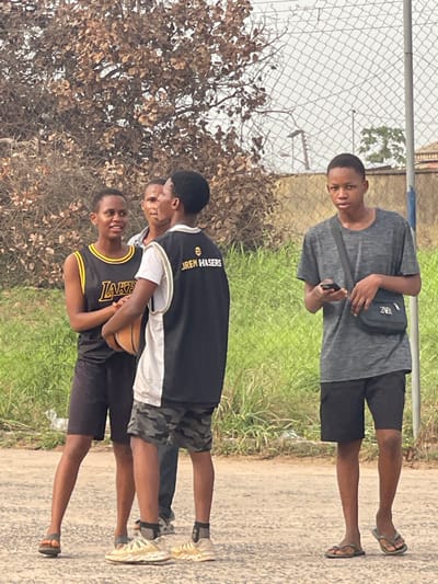 Four young men pose outdoors. The top individual sports a gray T-shirt, black shorts, and sandals, with a black bag slung across his shoulder. Below, three individuals stand clustered. One wears a Lakers jersey and holds a basketball. The second wears a black and white jersey with "Lauren Hasers" visible, and the third is partially obscured. They all wear shorts and shoes except for the top individual who wears sandals. The backdrop includes trees, grass, and a chain-link fence.