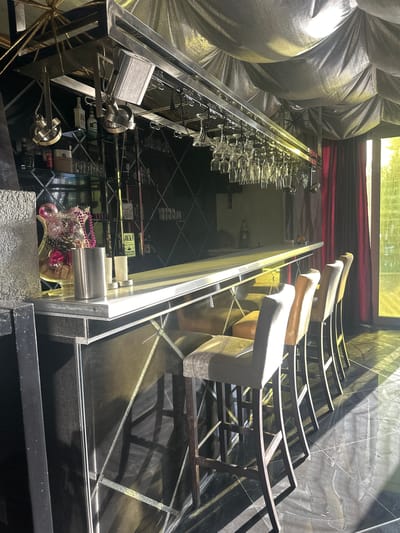 The image depicts a dimly lit bar interior with a long, polished counter. Several bar stools, upholstered in what appears to be light-colored fabric, are arranged in a row in front of the counter. Behind the counter, shelves are stocked with various bottles and glassware. A rack of wine glasses hangs from the ceiling above the counter. The ceiling is covered with a textured, reflective material, and lighting fixtures are visible. To the right, a set of dark red curtains hangs, and beyond them, a window allows sunlight to stream into the room, casting long shadows on the floor. The floor itself is made of dark, polished material with visible veins. On the left side of the counter, a glass vase contains decorative items, including what look like flowers and wine corks. The overall ambiance suggests an upscale or stylish establishment. There are no people visible in the image. The location is identified as Brca, Montenegro.