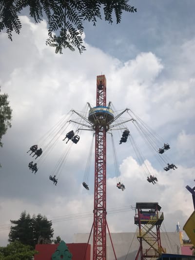 The image shows a red and white amusement park ride, a swing ride,  in operation.  There are 12 people on the ride. The ride is located in Batu, Indonesia at the coordinates provided (-7.885, 112.524). The ride is called the “Flying Fox.” The ride is in a park, with trees, cloudy skies, and a building in the background.