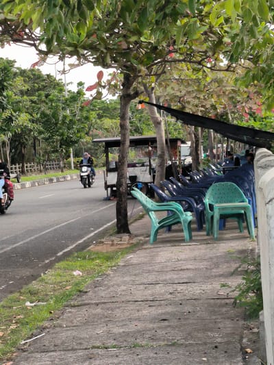 This image depicts a roadside scene with a focus on a sidewalk lined with numerous blue and green plastic chairs. In the background, a street is visible with a few people riding motorcycles. One motorcycle, ridden by a person wearing a hijab, is in the foreground, traveling on the road. Behind the chairs, a small food stall or cart can be seen, suggesting a potential gathering or dining area. Trees with lush green leaves and some red foliage overhang the scene, casting shadows. The weather appears overcast, and the time of day is difficult to determine but likely daytime. The setting seems to be a public area, possibly a tourist spot or a casual eatery, given the presence of the chairs and food stall. There is no visible text in the image.
