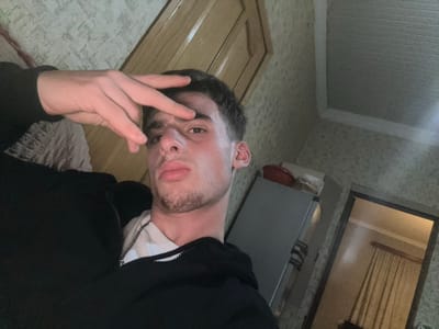 A young male subject is depicted in an indoor selfie, positioned in the lower-left to central frame, looking directly at the camera. He has short dark hair and light facial stubble, wearing a black zippered hoodie over a white t-shirt. His left hand is raised, forming a V-sign with the index and middle fingers, positioned over his forehead and partially obscuring his left eye.

The background shows a domestic interior. To the subject's right, a light-colored refrigerator is visible, topped with a red bowl and a lighter, undefined object. The walls are covered in a light-colored, patterned wallpaper. In the upper-left quadrant, a portion of a wooden door or cabinet is visible. A doorway on the far right opens into another room, revealing light-colored curtains. The ceiling is light-colored and appears textured.

The specific city of Durgena, Georgia, cannot be visually confirmed from the details present in the image.