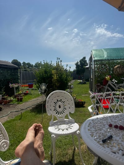 The image shows a sunny garden with white metal patio furniture. In the foreground, a person's bare feet are resting on the grass, with one foot crossed over the other. The furniture includes a table with decorative cutouts and a chair. The garden is green with a pathway winding through it. In the background, there is a greenhouse with green netting, some trees, and a fence. The sky is blue with scattered white clouds, indicating a pleasant day, likely late morning or early afternoon due to the strong sunlight casting shadows. The overall scene evokes a sense of relaxation and enjoyment of a summer's day.