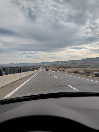 A tilted image showcases a highway stretching into the distance. On the left, a cloud-filled sky meets land featuring rows of buildings. The highway has a white dividing line and several vehicles can be seen in the distance. The right side of the image includes a portion of a vehicle interior, likely the dashboard and steering wheel, suggesting the photo was taken from inside a car.