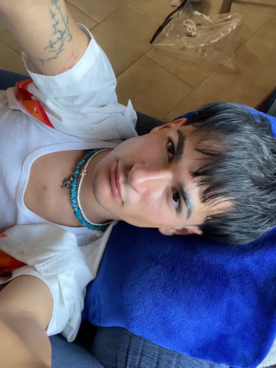 An upward-facing portrait captures a person lying down, looking directly into the camera, likely a selfie. The individual appears to be a young adult with dark, slightly messy hair that has some grey streaks, falling forward over the forehead. Their face has a neutral expression, with dark eyes and what appears to be sweat or moisture on their forehead and nose.
They are wearing a white ribbed tank top layered under an open white short-sleeved collared shirt. The inside lining of the collared shirt shows a pattern of orange and white fabric. Two necklaces adorn their neck: one made of blue-green beads with a silver-colored charm, and another with white beads and a small white shell-like charm. Visible on their left arm is a tattoo featuring dark, abstract, branch-like patterns and some faint pink or red markings.
The person's head rests on a vibrant blue, soft-textured pillow or towel. To the left, part of a dark blue, possibly denim, fabric is visible, suggesting they might be wearing jeans or sitting on a similar textured surface. The background on the right reveals a light-colored, possibly beige or brown, tiled floor. A clear plastic bag containing indistinct white objects is also seen on the floor. The setting appears to be indoors, giving a relaxed and casual atmosphere. No specific time of day or weather conditions are evident. There is no visible text in the image.
