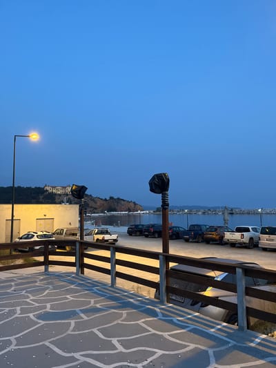 The image shows a view from a patio in Limenaria, Greece (latitude: 40.626, longitude: 24.575). The patio is made of gray stone tiles with white lines, and it is surrounded by a wooden railing. The patio faces a parking lot with a row of parked cars and trucks. There is a small building on the left, a hill in the background, and a body of water in the distance. The sky is clear blue and the sun is setting. There is a street light on the left and two more lights in the foreground. There are no people in the image.