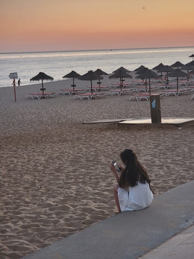 A woman with long, dark hair is seen from behind, sitting on a concrete ledge at a beach in Quarteira, Portugal, looking at her phone. She is wearing a white, eyelet dress. The beach is sandy, with rows of empty lounge chairs and thatched umbrellas in the background. The ocean stretches out to the horizon under a pastel-colored sky, indicating either dawn or dusk. The scene is peaceful and deserted except for the woman.