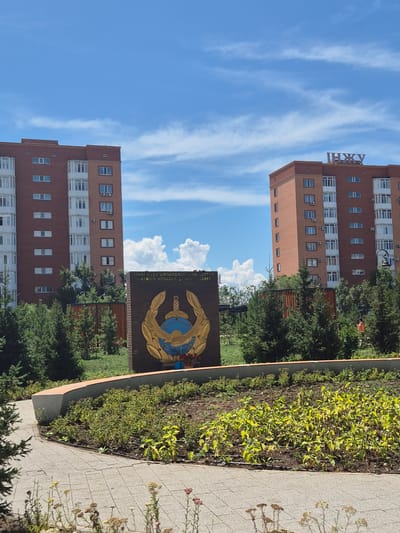 The image shows two tall, reddish-brown apartment buildings under a bright blue sky with scattered white clouds. The building on the left is larger and appears to be an older style, while the one on the right is slightly smaller and has a more modern appearance, featuring a sign with Cyrillic lettering that reads "ИНЖУ" on its roof. In the foreground, there is a monument or memorial made of dark brick, adorned with a gold emblem featuring a sword, a globe, and laurel leaves. The text on the memorial is partially visible and appears to be in a language using Latin script, possibly related to a military or governmental organization. The area around the memorial is landscaped with green bushes and trees, and a paved walkway made of square grey tiles is visible in the lower part of the frame. The overall scene depicts a public space in a city, likely a residential area given the apartment buildings. The location is identified as Kokshetau, Kazakhstan.
