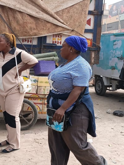 The image depicts an outdoor street scene in Fagge C, Nigeria, featuring two adult women and multiple vehicles. The ground consists of light brown, unpaved dirt.
On the left, a woman with braided yellow hair, pulled into a ponytail, stands facing left, with one hand on her hip. She wears a beige tracksuit-style outfit with black geometric patterns on the collar, shoulder, and lower leg. A white shoulder bag with gold-colored hardware hangs from her left shoulder. She wears black open-toed sandals.
Adjacent to the woman on the left is a wooden handcart with a spoked wheel, laden with numerous cardboard boxes, some marked "DEL." On top of the boxes sits a purple plastic basket containing several rolled green mats. Behind the cart and the left woman is the rear of a dark blue truck, partially covered by a tan canvas tarp. White lettering "PH" and "50 KM PH" is visible on the truck's side.
On the right, a second woman walks towards the right side of the frame, her head turned slightly left. She wears a blue headwrap, a blue and white horizontally striped short-sleeved t-shirt with a white graphic on the chest, and dark brown trousers. A dark blue garment is tied around her waist. She has a silver stud earring in her right ear and carries a black cross-body bag with a blue floral design on its front. Her footwear is partially visible and dark-colored.
In the background, beyond the right woman, a light blue commercial vehicle with "3 in 1" branding is visible. Distant structures include buildings with commercial signage, specifically "FABULOUS" and "MALENA."