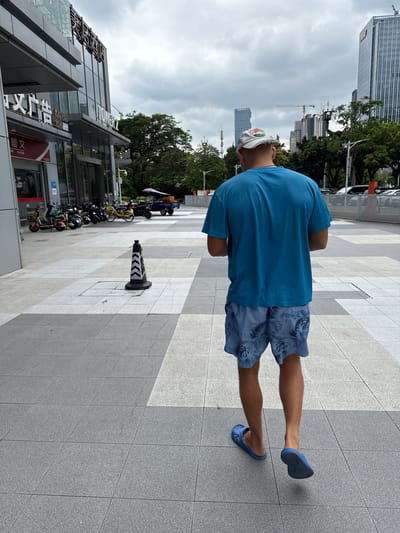 In Huangpu District, China, a man wearing a blue t-shirt and blue patterned shorts walks away from the camera on a paved walkway. He is also wearing blue flip-flops. In the background, a modern building with glass windows and signage is visible. To the left, several electric scooters are parked. Further in the distance, tall buildings and construction cranes indicate an urban setting. The sky is overcast. A black and white traffic cone is positioned on the walkway in the foreground.