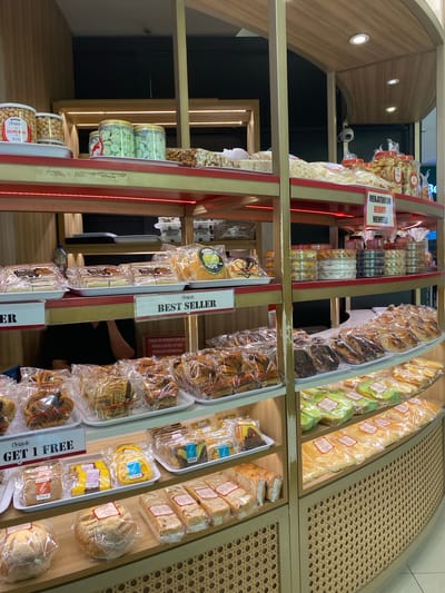 This is a photograph of a bakery display, likely taken in West Jakarta, Indonesia, given the Indonesian text on the signage. The bakery features multiple shelves laden with various baked goods and packaged snacks.
On the top shelf, several large plastic containers filled with what appear to be cookies or crackers are visible. One container, labeled "Dinamik Kacang Madu," contains honey peanuts. To the right, there are more containers of snacks, some with gold-colored lids.
The middle shelves display a variety of packaged baked goods. On the left, rows of individually wrapped cakes or pastries are arranged on white trays. A sign in the center reads "Dynamic BEST SELLER." To the right of this, more packaged baked goods are displayed, including some with prominent branding like "Dynamic."
The lower shelves showcase more baked items. On the left, there are packaged breads and pastries, with one prominent label indicating "GET 1 FREE." Further to the right, the shelves are filled with an assortment of cakes and breads, all individually wrapped in plastic. Some of these appear to be loaf cakes or layered pastries.
The overall environment suggests a retail setting, likely a supermarket or a dedicated bakery outlet. The display is well-lit, and the products are neatly arranged. The signage in Indonesian indicates the language of commerce and is common in retail establishments in Indonesia.