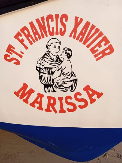 The image displays a close-up of the side of a boat, resting on sandy ground, indicative of a beach or shoreline in Arambol, India. The boat's hull is painted white on the upper section and a deep blue below.

Dominating the white section of the hull is a prominent graphic featuring a black-and-white illustration and red text. The illustration depicts an adult male figure, presumably St. Francis Xavier, in traditional religious robes, gently holding a child, likely the Infant Jesus. The adult figure wears a serene expression and holds what appears to be flowers in one hand.

Arched above the illustration in bold, red capital letters is the text "ST. FRANCIS XAVIER". Below the illustration, also arched in bold, red capital letters, is the name "MARISSA". Both sets of text have a slightly textured, possibly hand-painted or stenciled, appearance.

The boat's blue lower hull is partially visible at the bottom of the frame, with light brown sand underneath, suggesting the vessel is either beached or docked close to the shore. The bright, even lighting indicates it is daytime. The presence of the religious figures and names suggests the boat carries a dedication or blessing, a common practice for vessels in coastal communities.