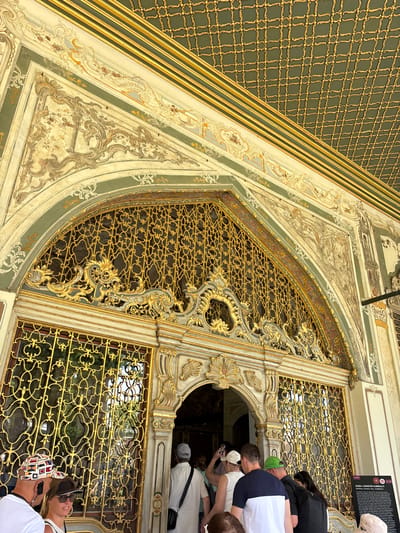 The image shows a detail of the ornate architecture of a building, likely a palace or historical site, in Istanbul, Turkey. The primary subjects are the intricate decorative elements of the building's facade, featuring a large, gold-colored metal latticework window or screen with elaborate scrolling designs. This latticework is set within a richly ornamented architectural frame, with painted and gilded stucco work in muted greens, creams, and golds depicting floral motifs and swirling patterns. A section of wall to the left features a dark green textured surface with a repeating geometric gold pattern.
Several people are visible, appearing as tourists observing the architecture. They are mostly seen from the back or side, with some looking at their phones. One person in the foreground is wearing a white top and a patterned baseball cap. Another person in the background wears a dark top and a white cap. The people suggest the activity of visiting a tourist attraction.
The time of day appears to be daytime, with the lighting suggesting a bright, possibly sunny day, casting some shadows. The overall impression is one of historical grandeur and detailed craftsmanship. A black sign with white text is partially visible in the upper right corner, likely providing information about the site, but the text is too small to be legible. The location context of Istanbul, Turkey, points to a significant cultural or historical landmark.