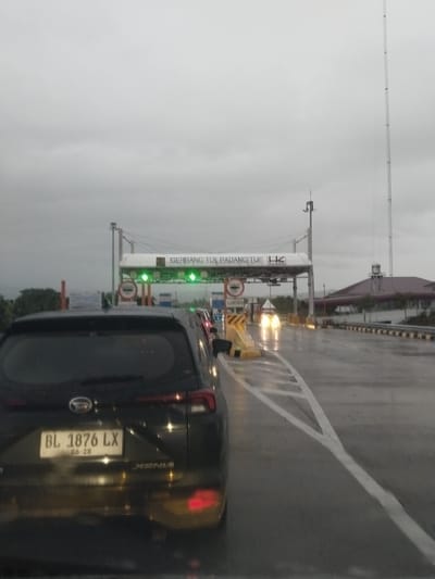 The image captures a view from inside a vehicle at a toll gate on a cloudy, rainy day. The vehicle's rear, including its license plate "BL 1876 LX" and the "XENIA" model name, is prominently in the foreground. The toll gate structure, labeled "GERBANG TOL PADANGTIJI", is visible ahead, with green lights indicating open lanes. Several other vehicles are queuing at the toll booths. The road surface is wet, reflecting the lights of the vehicles and the toll gate. A tall communication tower stands in the background to the right, and a building with a distinctive roof structure is also visible. The environment appears to be a highway or main road. The location is identified as Pante Cermen, Indonesia.