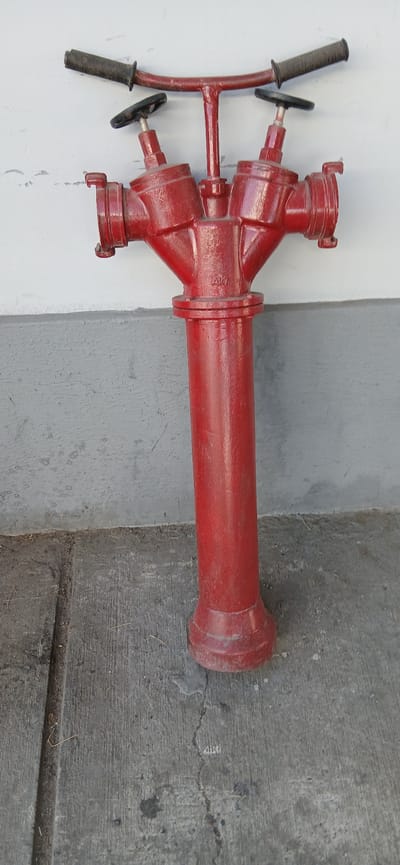 A red fire hydrant stands vertically against a white and grey wall, with a concrete ground in the foreground. The hydrant has a central vertical pipe with a flared base, topped by a triangular section with two spouts on either side. These spouts have connecting mechanisms with caps and circular valve handles. A horizontal bar with two black grips is attached to the top of the hydrant, possibly for easy operation. The hydrant appears to be made of metal and shows some signs of wear and tear, with dust and minor scuffs. The wall behind it is painted white at the top and grey at the bottom, with a textured surface. The concrete ground is cracked and shows some debris. The scene is well-lit, suggesting it is daytime. The location is identified as Beshkurgan, Uzbekistan.