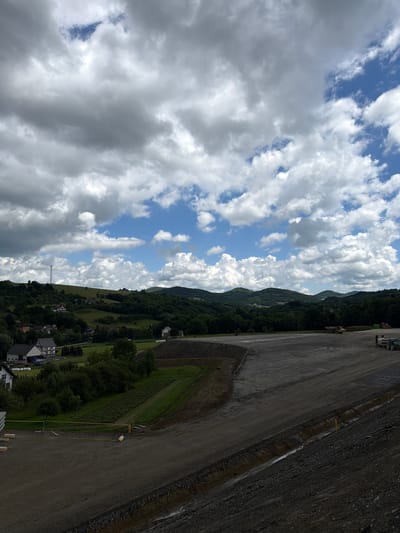 The image depicts a large, partially constructed area with a gravel surface, likely a landfill or construction site, located in the city of Stróża, Poland. The foreground is dominated by this graded, dark gray gravel expanse, with some rocky debris visible along the lower right edge. A yellow caution tape is strung across a portion of the area, indicating a boundary or hazard. Further back, green fields and trees lead up to rolling hills covered in dense green foliage. Several houses are scattered across the landscape, some with white siding and dark roofs, appearing to be residential dwellings. In the middle ground, a section of tilled earth or cultivated land with rows is visible. On the right side of the constructed area, heavy machinery is present, including a yellow dump truck, a green tractor, and another vehicle that appears to be a cement mixer. A tall, lattice-style tower stands on a distant hill to the left. The sky above is filled with dramatic, heavy clouds interspersed with patches of blue sky, suggesting a partly cloudy day.