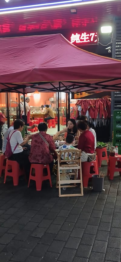 A group of people are dining at an outdoor restaurant under a red canopy. There are at least seven individuals visible, including adults and a young child in a high chair. The dining area is set up on a paved street. In the background, there is a building with illuminated red signage. Several red plastic stools are used as seating. Food and drinks are present on the table. The scene appears to be in the Jiulongpo District of China, as indicated by the context provided. The atmosphere suggests a casual evening meal.