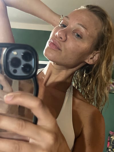 A close-up, low-angle, upside-down selfie shows a young woman with blonde, wavy hair. She is wearing a light-colored swimsuit or tank top. Her face is turned towards the camera, and her eyes are looking slightly to the side. She has on lip gloss and her tongue is slightly sticking out. The background is a green wall. The image appears to be taken indoors with artificial lighting, possibly in the evening or at night. The focus is on the woman's face and upper body, with her arm and hand holding a phone, which is capturing the reflection of her face. The scene is casual and personal, suggesting the photo was taken for personal use or social media.