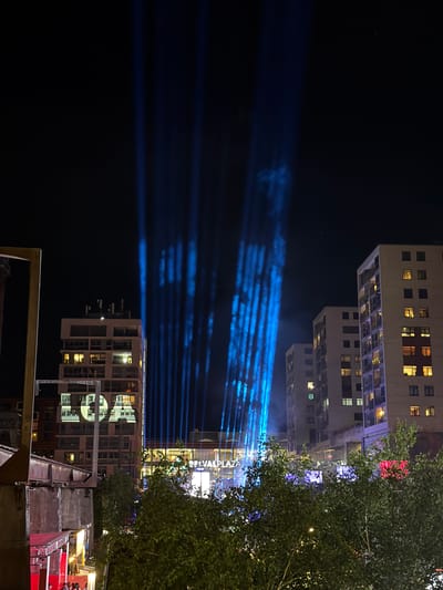 The image depicts a night scene in Esch-sur-Alzette, Luxembourg, with a view of Belval Plaza. Multiple buildings with illuminated windows are visible, some of which are partially obscured by tree foliage. Light beams emanate from the area of Belval Plaza, projecting horizontally across the dark sky. A foreground includes metallic structures and hints of red surfaces, suggesting elements of urban architecture or temporary installations.