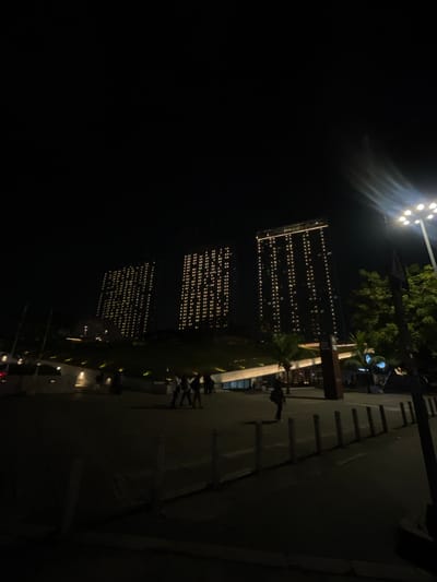 The image depicts a nighttime scene in the city of Central Jakarta, Indonesia, likely an urban development or waterfront area. Three tall buildings are visible in the background, illuminated by rows of lights on their facades, suggesting they are residential or commercial towers. The buildings are dark in color and appear to be modern high-rises.
In the foreground, several people are walking and standing. They are silhouetted against the illuminated buildings and surrounding lights, making individual features indistinct. Some individuals appear to be gathered in small groups, while others walk alone. The ground is paved, and a line of bollards or small posts runs along the edge of a walkway.
The environment is dimly lit, with artificial lighting from the buildings, streetlights, and possibly the entrance to a building or establishment. There are trees and palm trees visible, indicating a tropical or sub-tropical climate. The overall impression is of a public space, possibly a promenade or plaza, adjacent to modern urban architecture, active at night. The lighting creates a dramatic and somewhat atmospheric scene.