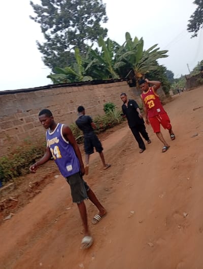 Four young men stand on a dirt road near a brick wall. One wears a Lakers jersey and cut-off shorts with sandals, another a black shirt and shorts. A third man in a black suit holds a phone, and the fourth wears a Cleveland Cavaliers jersey and red shorts. Banana trees and other foliage grow nearby. The sky is overcast.
