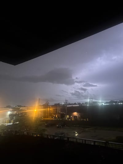 This is a nighttime, medium-distance, eye-level shot of a city street in Des Plaines, United States, during a thunderstorm. The foreground is dominated by a dark, overhanging structure, possibly a balcony or roof. Beyond this, the middle ground reveals buildings with illuminated windows and a street with wet pavement reflecting the light. Several cars are parked on the street. The background features a dramatic sky with dark, heavy storm clouds and visible lightning strikes. The overall atmosphere is dark and turbulent, indicative of severe weather.
