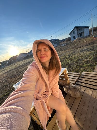 A young woman is captured in a selfie, relaxing outdoors at what appears to be sunset in Gornyi (Dvurechenskoe S/P), Russia. She has long, light brown hair and is wearing a cozy pink hooded robe, which drapes around her. A gentle smile graces her face, conveying a sense of contentment. Her legs are crossed, and she is wearing white, fuzzy slippers, suggesting a comfortable and relaxed posture. She appears to be seated on or near wooden pallets.

To the right of the woman and slightly behind her, a fluffy grey cat is visible, partially obscured, resting among more wooden pallets. The immediate ground in the scene consists of dry, sparse grass or dirt.

The background reveals a clear blue sky, illuminated by the low-hanging sun on the right side of the frame, indicating either late afternoon or sunset. On the left side of the background stands a large, grey, two-story house, which appears to be unfinished or newly constructed, featuring dark windows. Further in the distance, another smaller building is visible. The landscape is also marked by power lines and utility poles, suggesting a rural or developing residential area. There is no visible text in the image.