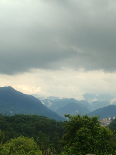 This media file depicts a hazy, mountainous landscape near Smolyan, Bulgaria. The scene is dominated by layers of blue-green mountains receding into the distance, shrouded in a light mist or fog that softens their outlines. A thick, dark gray cloud layer hangs low in the sky, obscuring the sun and creating a muted, atmospheric light. Hints of pale yellow light break through the clouds in the middle distance, illuminating the upper portions of some peaks.

In the foreground, a dense, dark green forest occupies the lower portion of the frame. The trees are lush and full, with some taller, more individual trees standing out. To the right, a cluster of buildings, appearing to be a small town or village with red-tiled roofs, is nestled on the side of a mountain in the far distance, partially obscured by the atmospheric conditions.

The overall impression is one of a tranquil, natural setting on a cloudy or overcast day. The weather appears to be humid and perhaps damp, given the mist. The time of day is not definitively clear, but the diffused light suggests it could be morning or late afternoon. No people, objects, or text are clearly visible in the image. The mood is serene and somewhat subdued due to the overcast weather.
