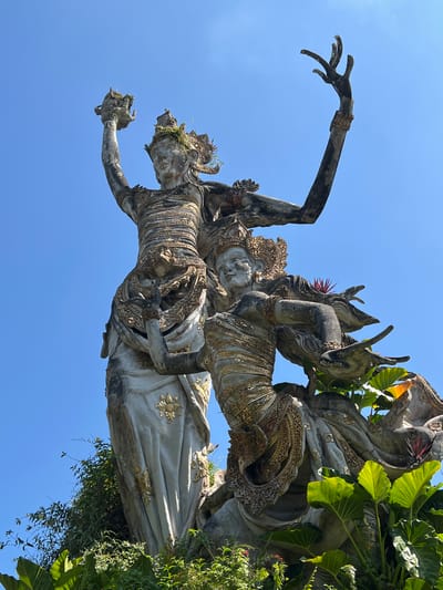 The image shows a stone sculpture in Kedewatan, Indonesia. Two figures are entwined; the top figure is smaller and appears to be riding on the larger figure's back. Both figures are adorned with elaborate headdresses and decorative garments. The sculpture exhibits some weathering and is partially covered in moss and foliage. The background is a clear blue sky. To the right, green leaves are partially covering the sculpture.