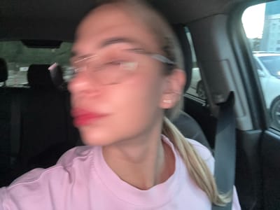 The image is a blurry, upside-down selfie taken inside a vehicle. The subject is a person with blonde hair, wearing a pink sweatshirt and glasses. Their face is largely out of focus, but their red lipstick is visible. The interior of the car is dark, with some of the car's interior features visible, including a seatbelt buckle. The background is also blurred, but appears to be the exterior of the car, possibly at night or in a dimly lit area. There is no visible text in the image. The overall impression is of a casual, candid photograph.
