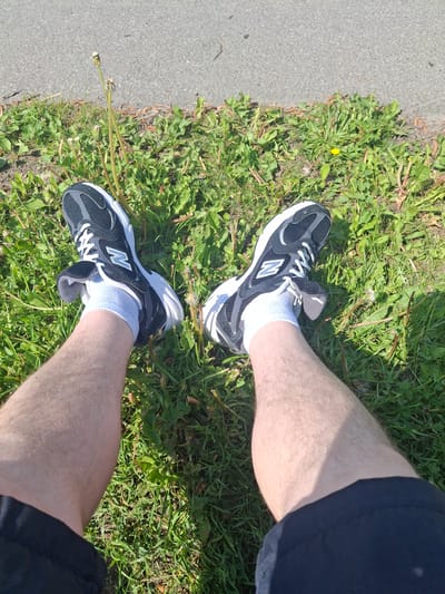 This media file captures a top-down view of a person's legs and feet resting on a grassy verge next to an asphalt path in Sillamäe linn, Estonia. The subject's bare legs are light-skinned and partially visible, extending away from the viewer. The person is wearing black and white New Balance sneakers, each featuring a distinct white "N" logo on the side, paired with white ankle socks.
The setting is outdoors on a bright, sunny day, evidenced by the strong sunlight and visible shadows cast on the grass and subtly on the legs. The ground consists of a textured grey asphalt path on the left, bordering a lush green area of grass and various weeds, including dandelions, some with small yellow blossoms and others in their white, seeding stage. The overall scene suggests a moment of rest or pause in an open, natural-urban environment, possibly a park or a pathway. No specific activity or emotion is discernible beyond the static pose of the legs and feet.