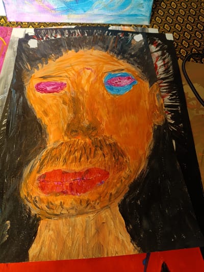 A large, abstract, vertical painting depicts a distorted, orange face with black hair. The face has prominent, bright pink lips and two distinct eye areas, one with pink and the other with bright blue. A brown mustache is visible above the lips. The background of the painting is black. The artwork is placed on a flat surface, possibly a table, with other artistic materials visible in the background, including parts of other paintings and what appears to be fabric with a patterned design. The image is cropped, showing a portion of the artwork and its surrounding environment. The painting is not located in Ban Patan, Thailand, nor does the image contain any contextual information to suggest this location.