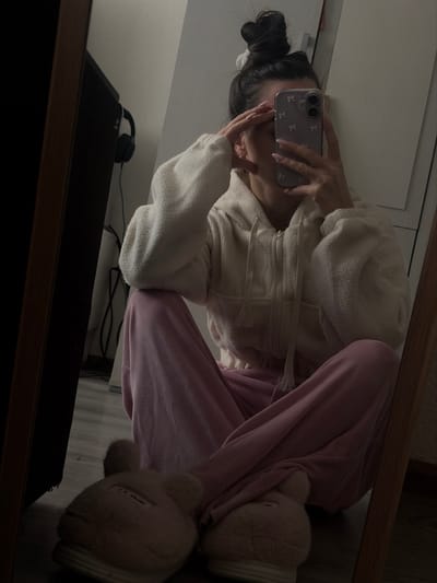 The image depicts an indoor scene, consistent with a private residence or room, located within Novosaratovka, Russia. A single individual, appearing female, is seated on a light-colored wooden floor, positioned to take a mirror selfie. The subject is wearing a light cream-colored, textured, full-zip hooded jacket with drawstrings, and pink velour or fleece sweatpants. On their feet are light pink, fuzzy slippers designed to resemble animal heads with prominent ears. The individual's dark hair is pulled into a high bun, secured by a white scrunchie. A light purple smartphone, featuring a clear case with a white floral or bow pattern, is held in the right hand directly in front of the subject's face, obscuring facial features. The left hand is positioned with fingers near the forehead, partially covering the upper part of the face. The fingernails are painted a light, neutral color. The background, visible in the mirror's reflection, shows a dark-framed object to the left, possibly a screen or piece of furniture, with black over-ear headphones hanging on it. To the right, a light-colored wall or wardrobe is visible. The scene is illuminated by subdued ambient lighting.