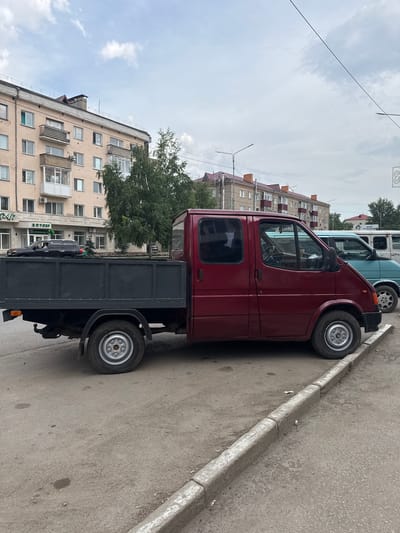 The image depicts a street scene in Kokshetau, Kazakhstan. A red Ford Transit van, modified with a flatbed truck body, is parked on the side of a paved road. The van is the primary subject, positioned in the center of the frame, facing to the right. Behind it, two other vans are partially visible: a light blue one to the immediate right, and a white one further back. The background features a multi-story apartment building with beige and pink facades and balconies, characteristic of Soviet-era architecture. A street with a concrete curb runs in front of the parked vehicles. A large green tree is situated between the apartment building and the van. The sky is overcast with clouds. No people are visible in the image, and the scene appears static, with no discernable actions taking place. The overall environment suggests a typical urban street in a post-Soviet city.
